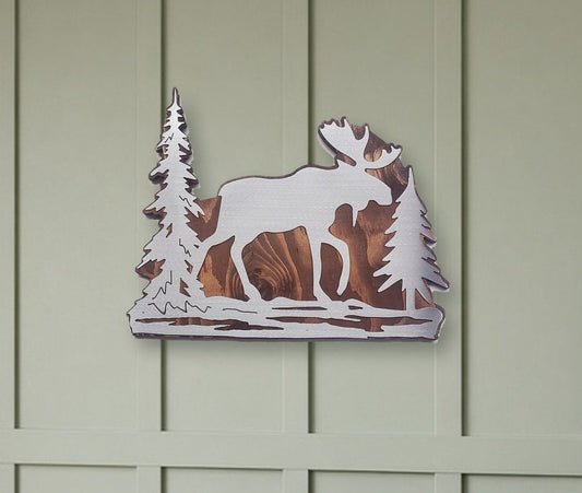 A rustic metal wall art piece depicting a moose and pine trees, mounted on a sage green paneled wall above a wooden bench with a wicker basket and brown boots.