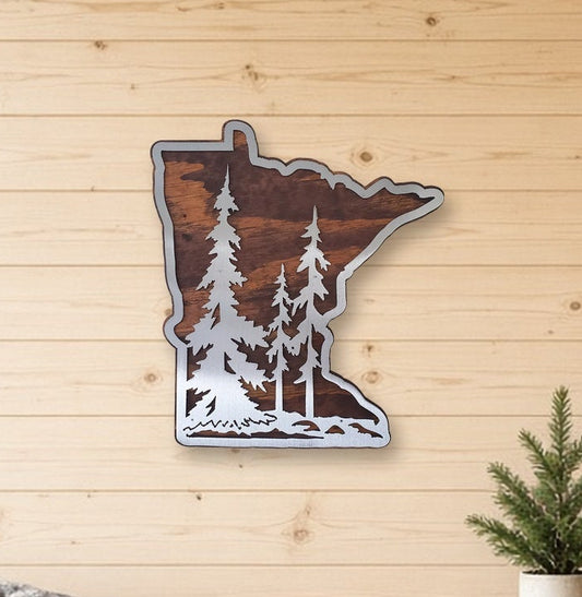 A decorative metal wall sign shaped like the state of Minnesota, featuring a brown forest silhouette with white trees, displayed on a light wood-paneled wall above a wooden bench in a rustic room setting.
