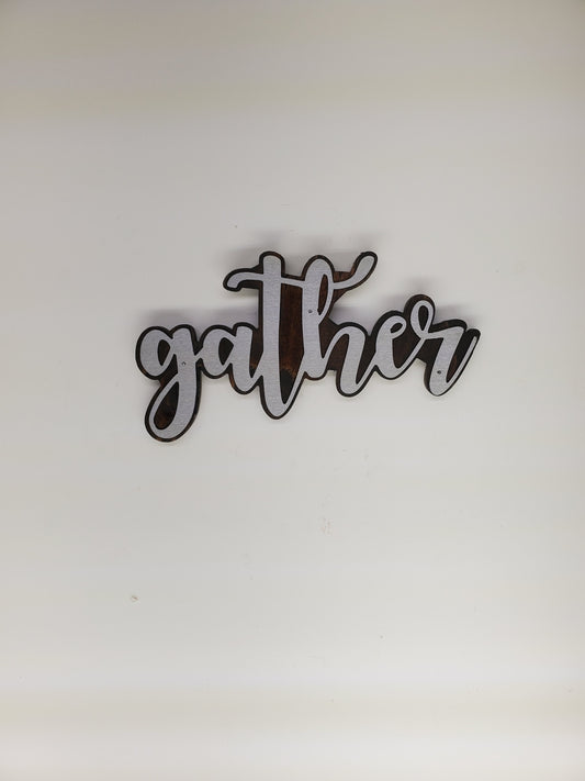 The metal art is cutout in the shape of the word "Gather", adding a touch of elegance and warmth to any room