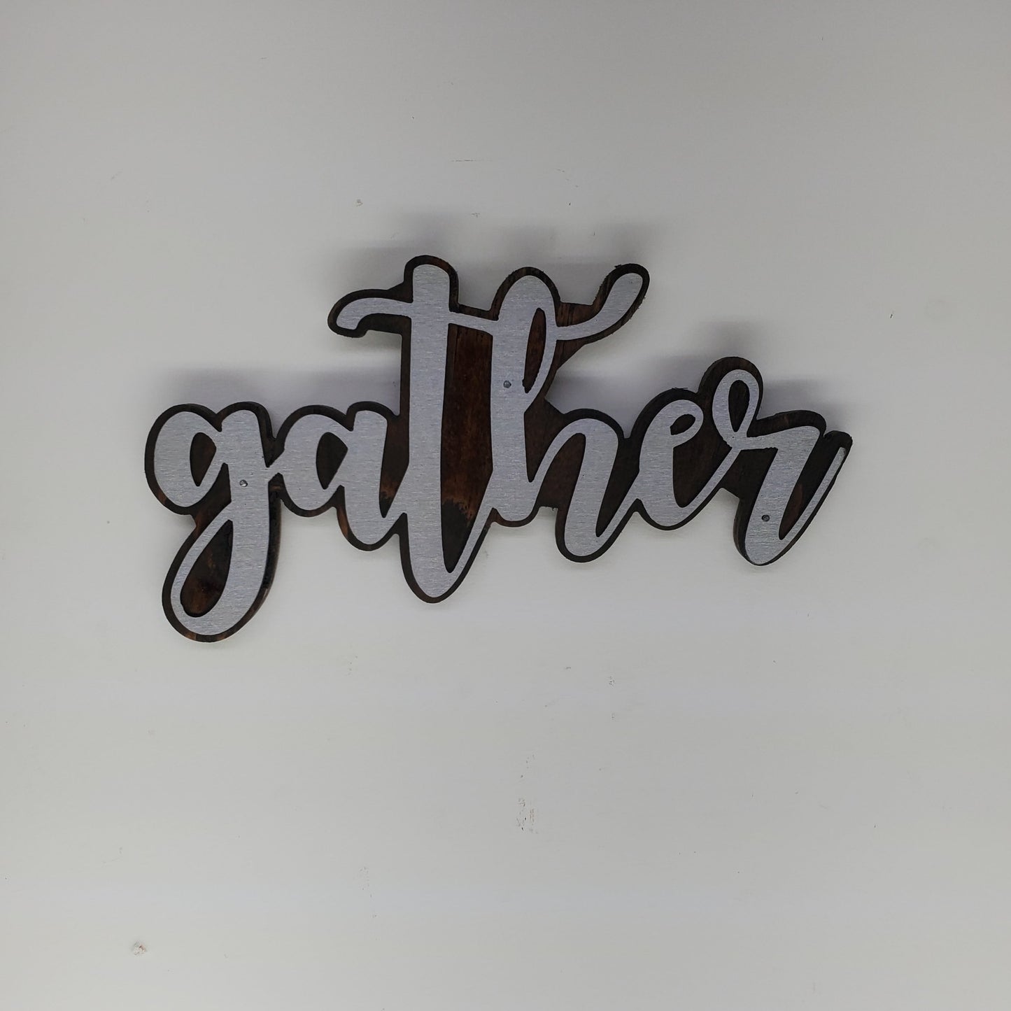 The metal art is cutout in the shape of the word "Gather", adding a touch of elegance and warmth to any room