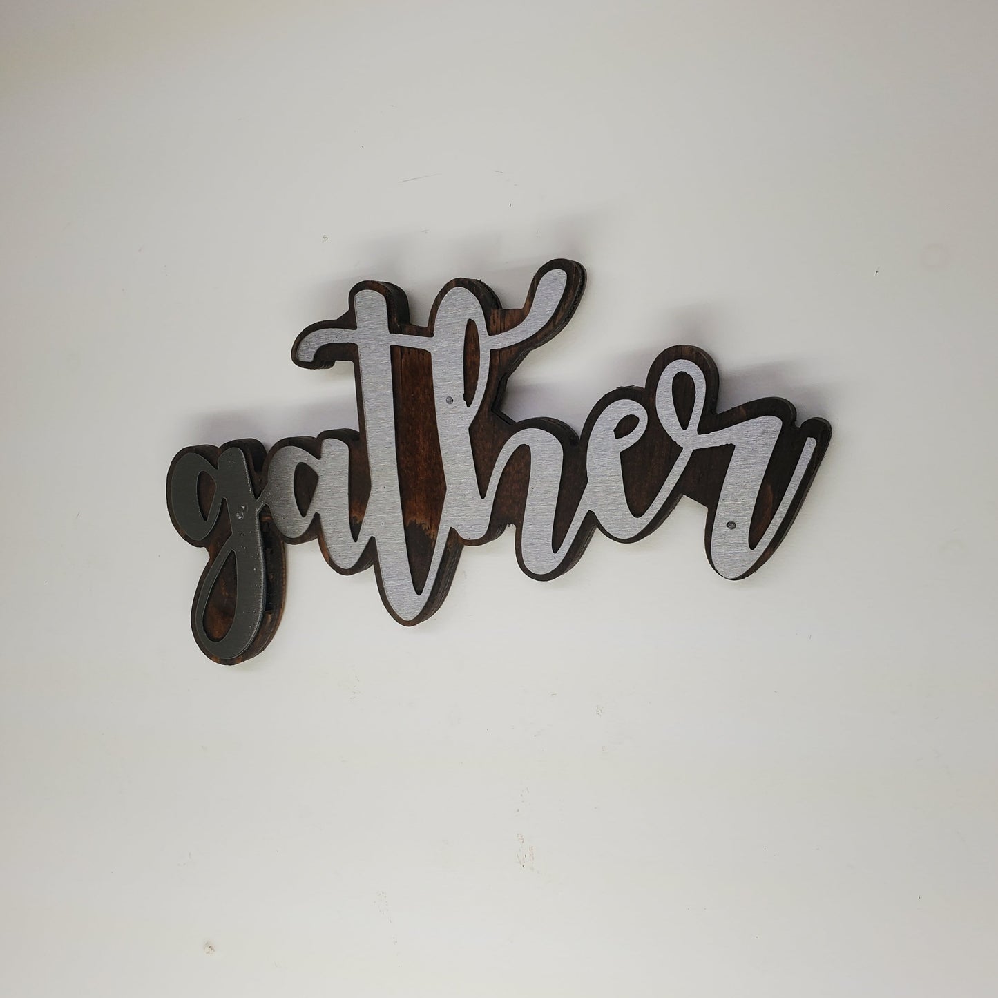 The metal art is cutout in the shape of the word "Gather", adding a touch of elegance and warmth to any room