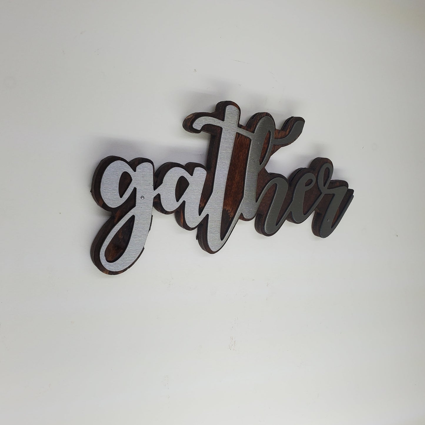 The metal art is cutout in the shape of the word "Gather", adding a touch of elegance and warmth to any room