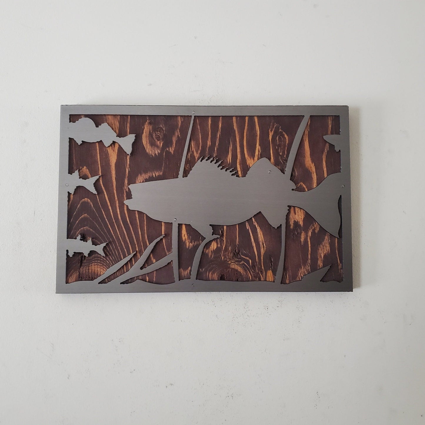 Metal art cut out of a walleye fish swimming in a natural habitat, mounted on a stained wooden background. The details of the fish and the surrounding nature are captured perfectly. The art work has an installed hanger on the back for easy hanging