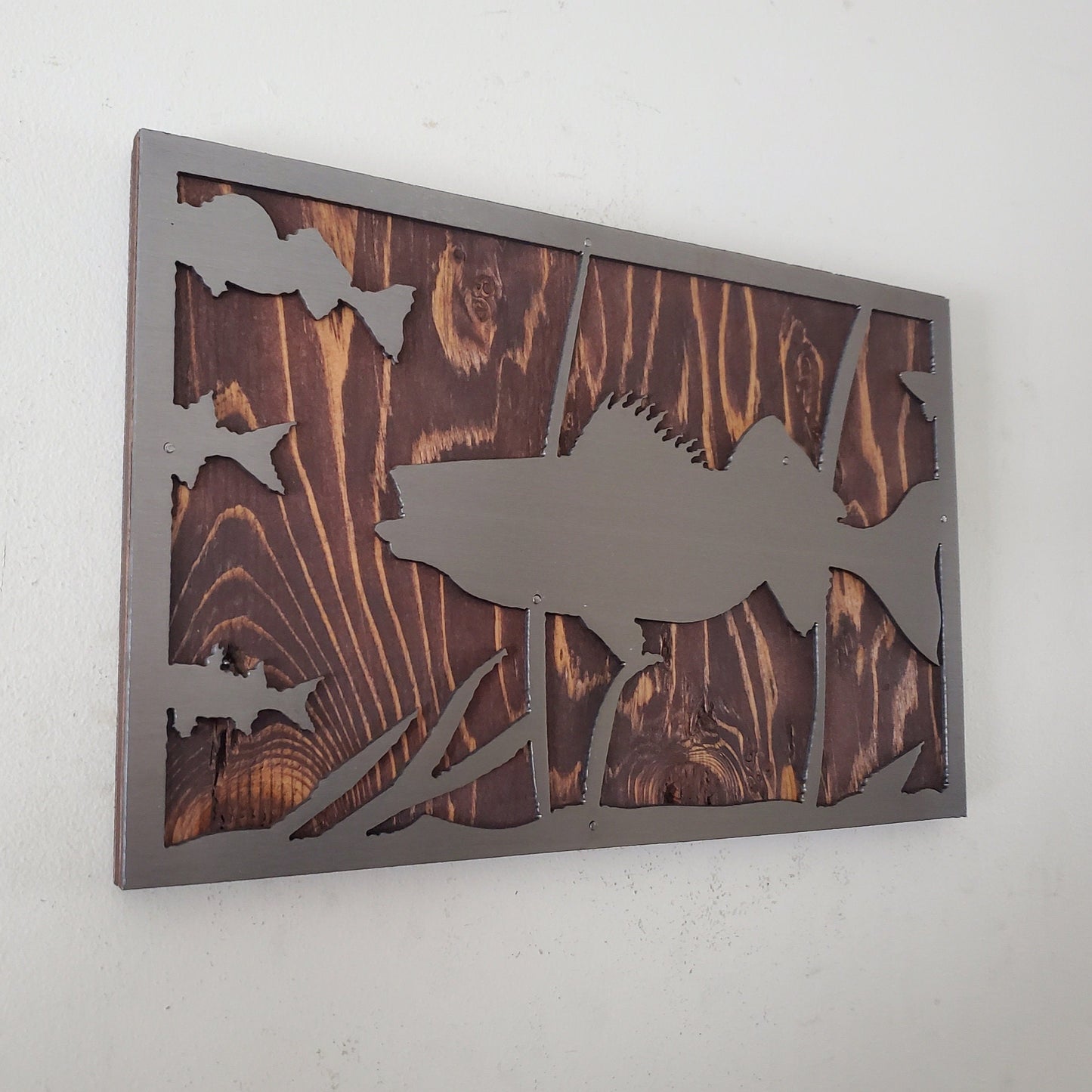 Metal art cut out of a walleye fish swimming in a natural habitat, mounted on a stained wooden background. The details of the fish and the surrounding nature are captured perfectly. The art work has an installed hanger on the back for easy hanging