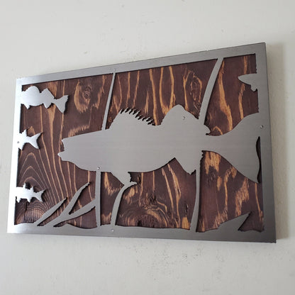 Metal art cut out of a walleye fish swimming in a natural habitat, mounted on a stained wooden background. The details of the fish and the surrounding nature are captured perfectly. The art work has an installed hanger on the back for easy hanging