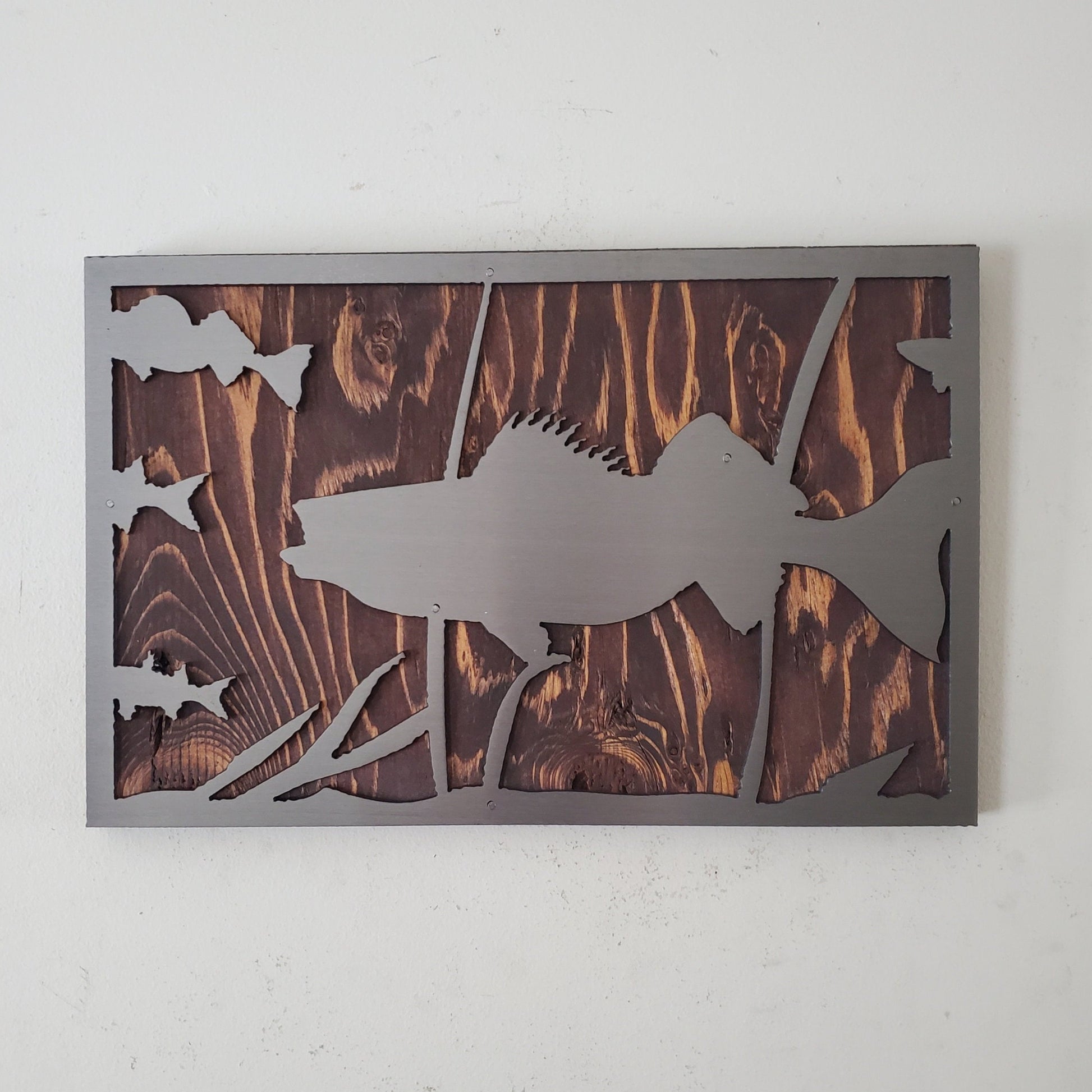 Metal art cut out of a walleye fish swimming in a natural habitat, mounted on a stained wooden background. The details of the fish and the surrounding nature are captured perfectly. The art work has an installed hanger on the back for easy hanging